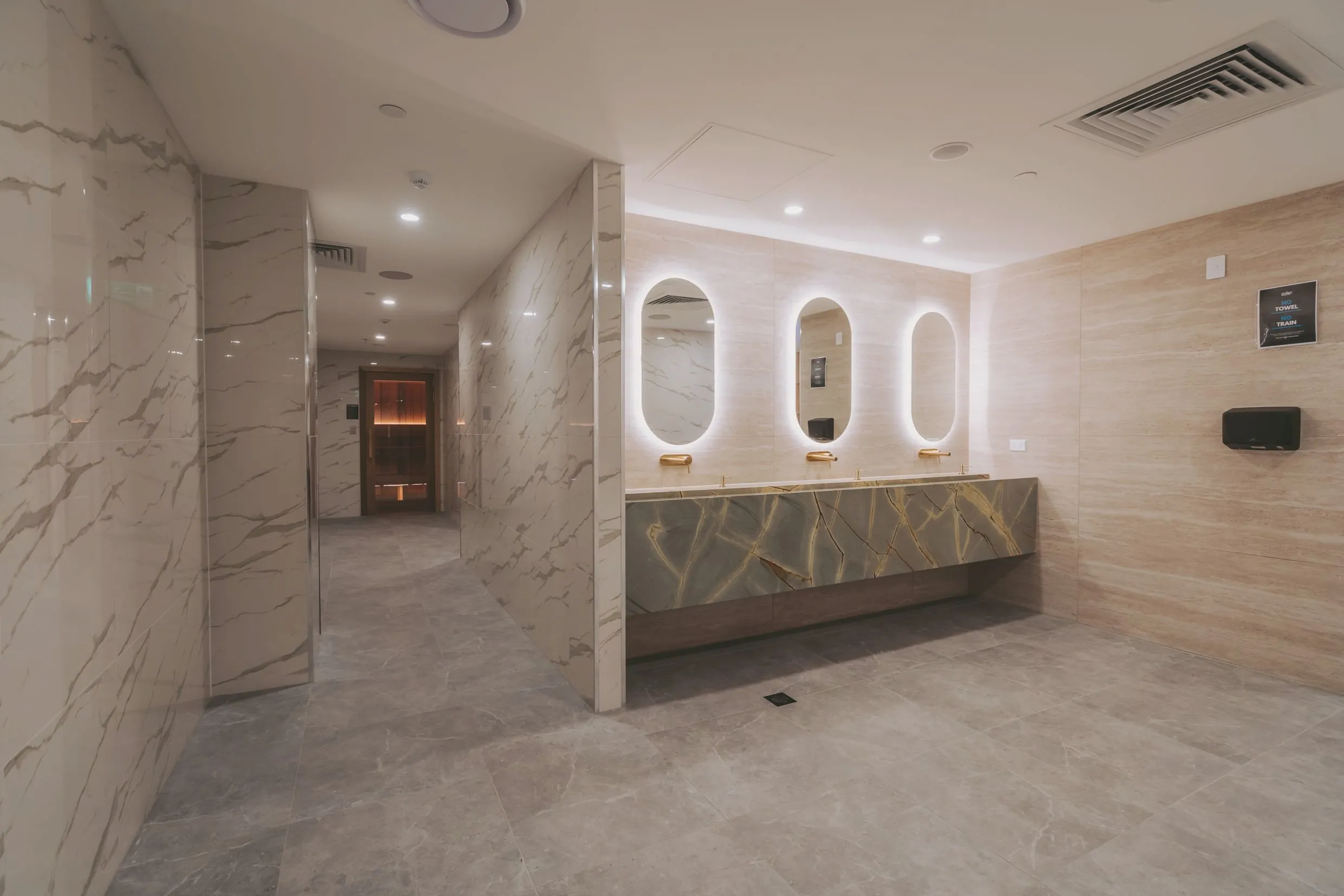 Marble-lined changing rooms with backlit oval mirrors, brass tapware and a stone vanity; the sauna door glows at the end of the corridor.