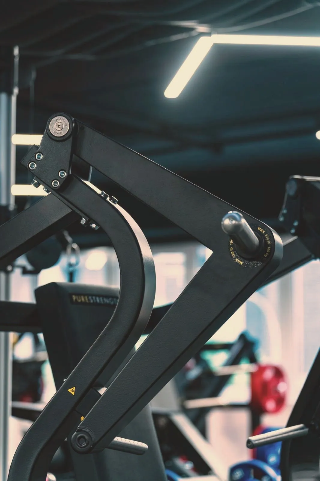 The pressing arm of a plate-loaded chest press in close-up, its machined pivot and plate horn against the blurred training floor.