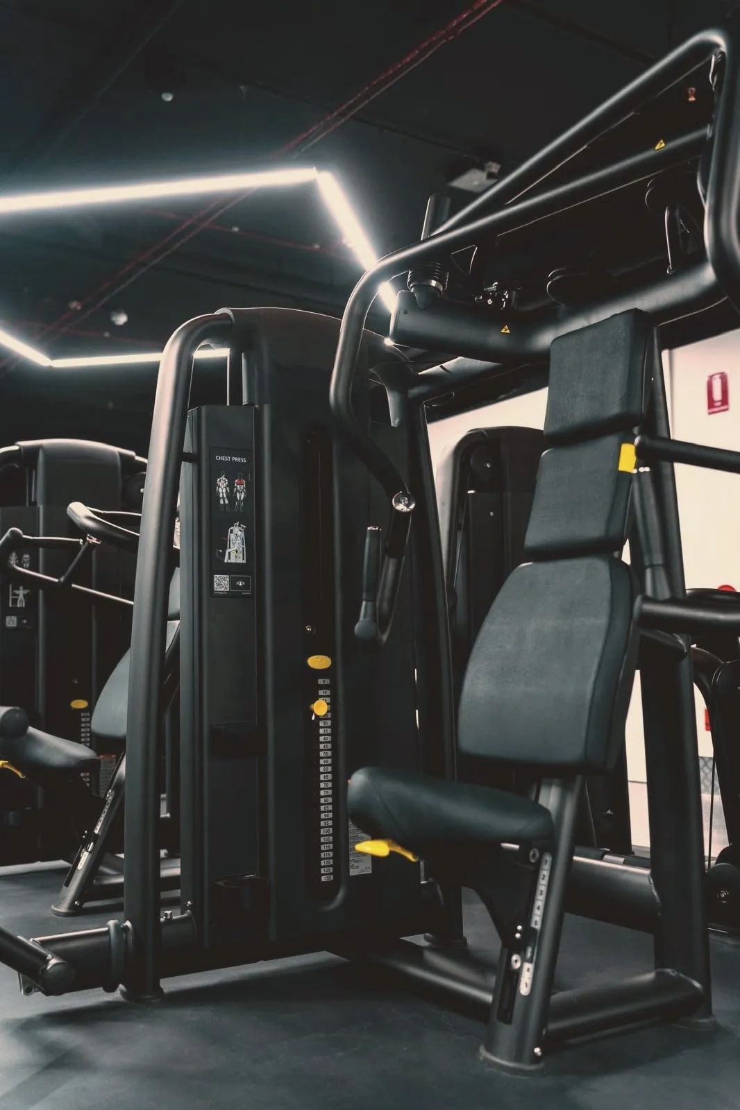 A row of selectorised chest-press machines in matt black, weight stacks racked beneath angled lines of light.
