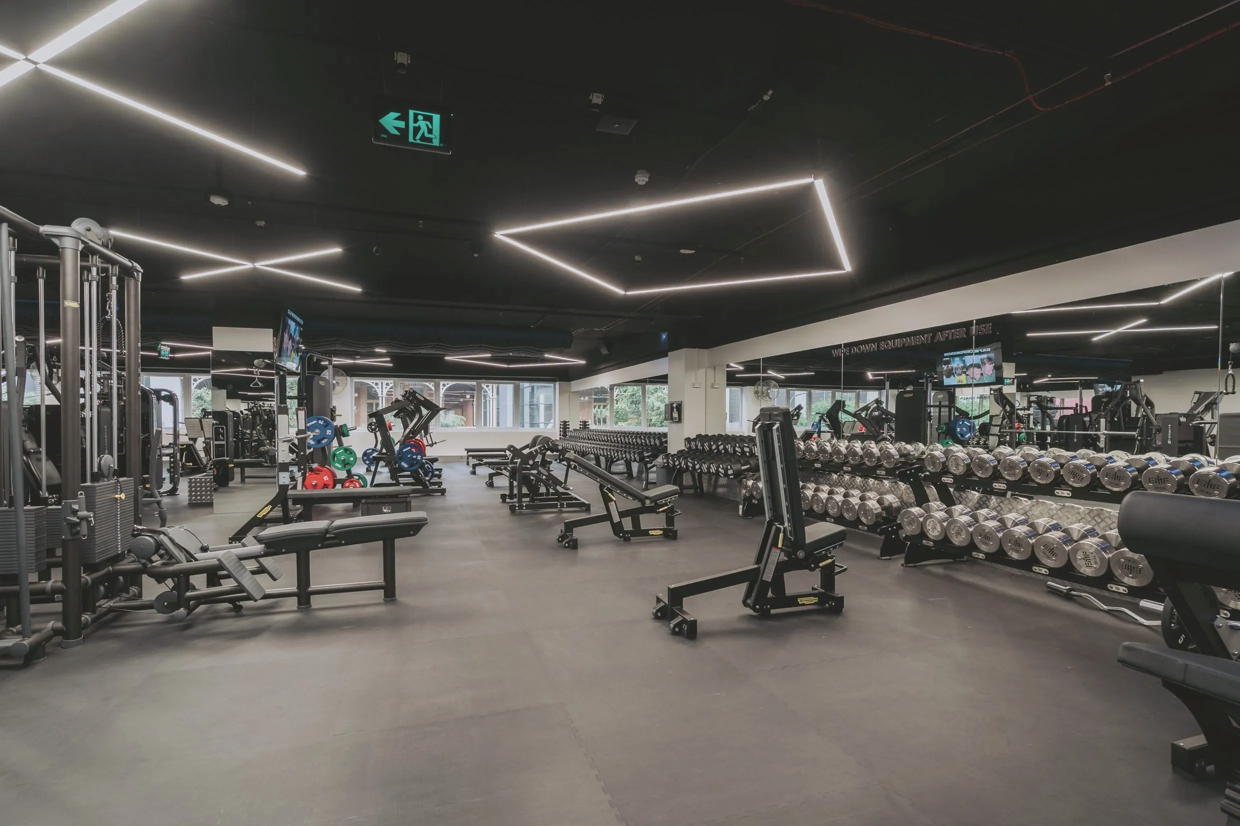 The free-weights floor: twin tiers of chrome dumbbells, adjustable benches and cable stations under a black ceiling, daylight from the street-side windows.