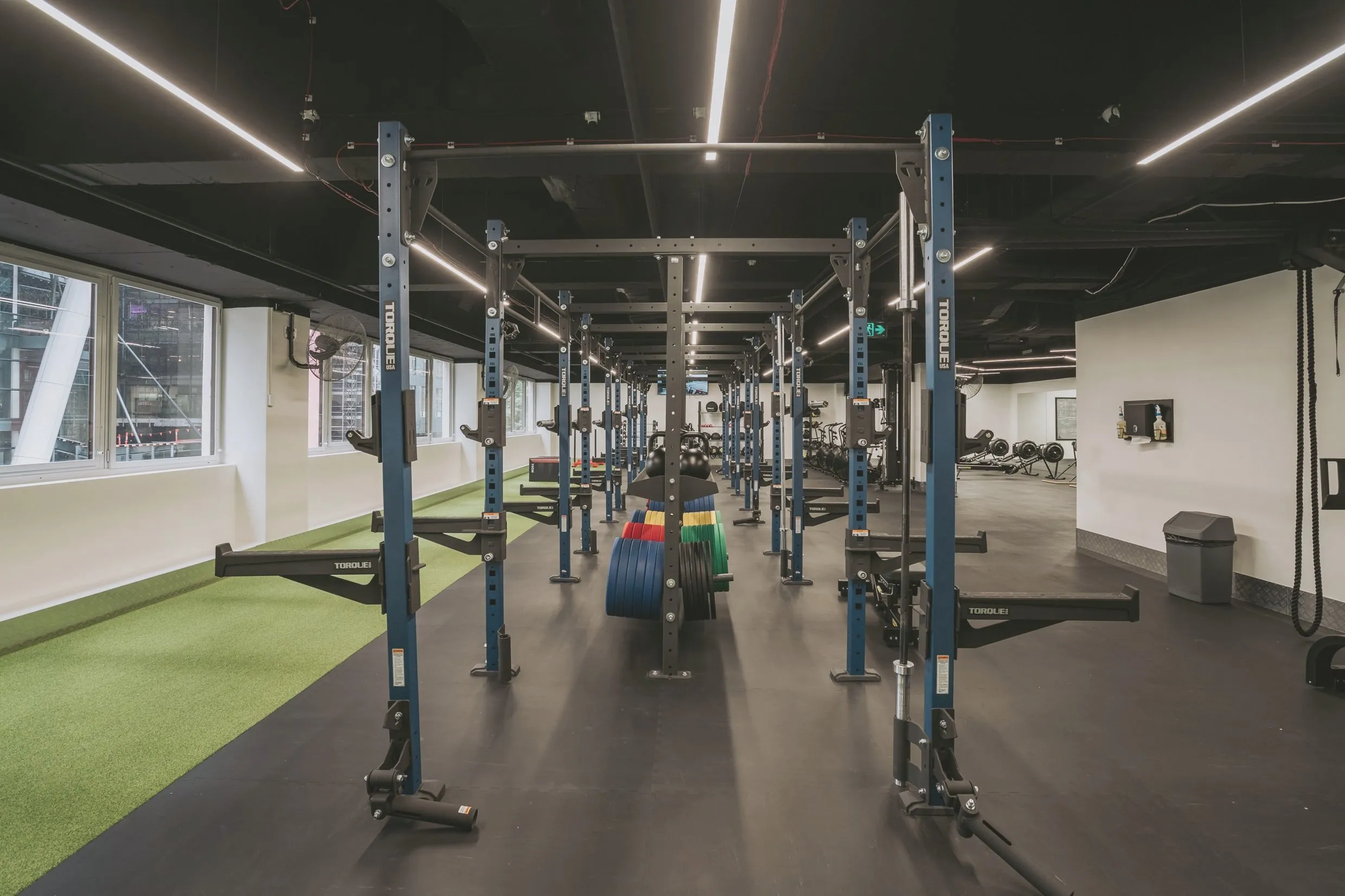 Paired racks down the centre of the performance zone, bumper plates staged between, a turf strip to one side and ergometers beyond.