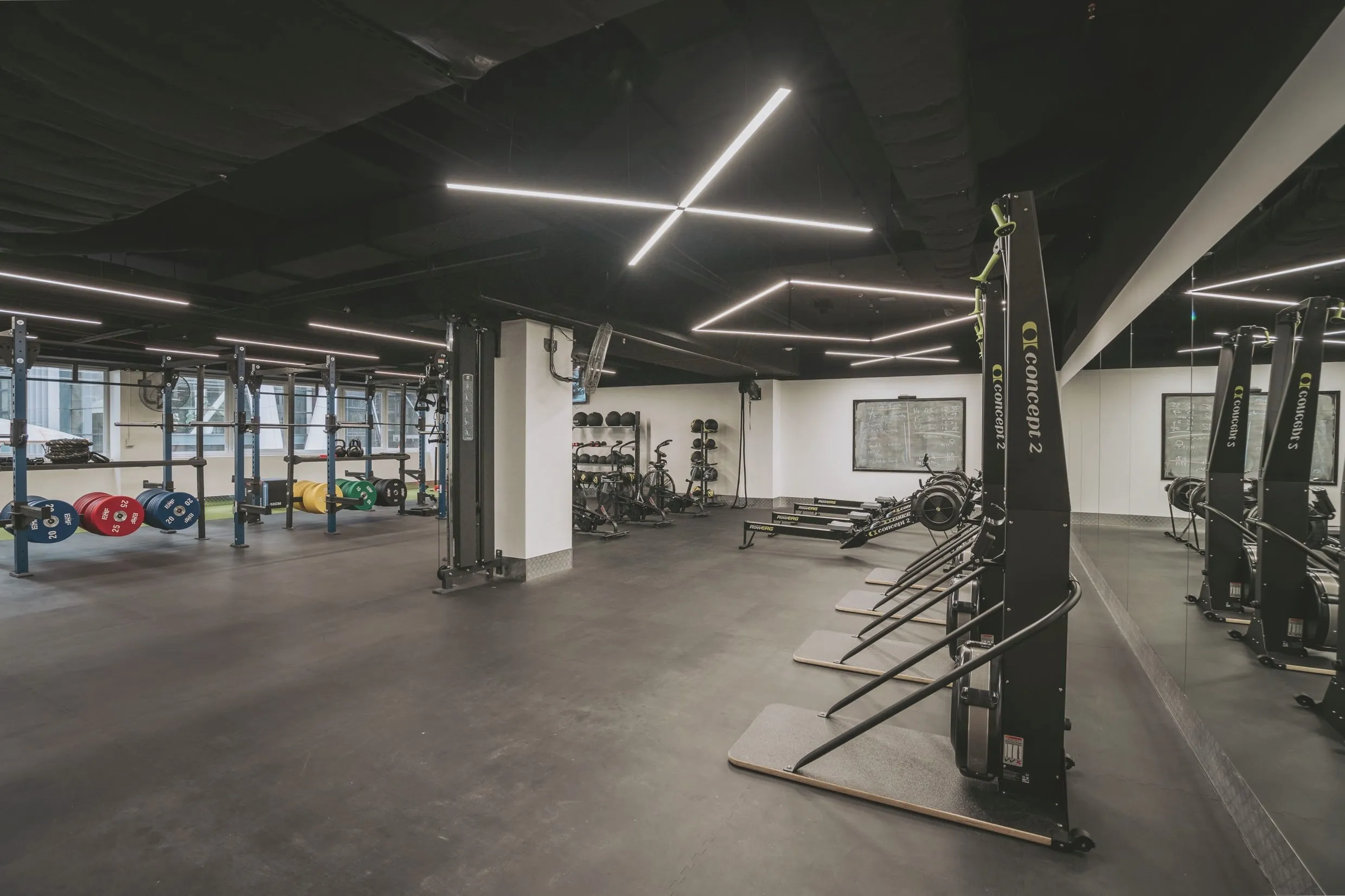 The performance zone: racks and bumper plates beside a strip of turf, with ski and rowing ergometers along the mirrored wall and a chalkboard of the day’s work.