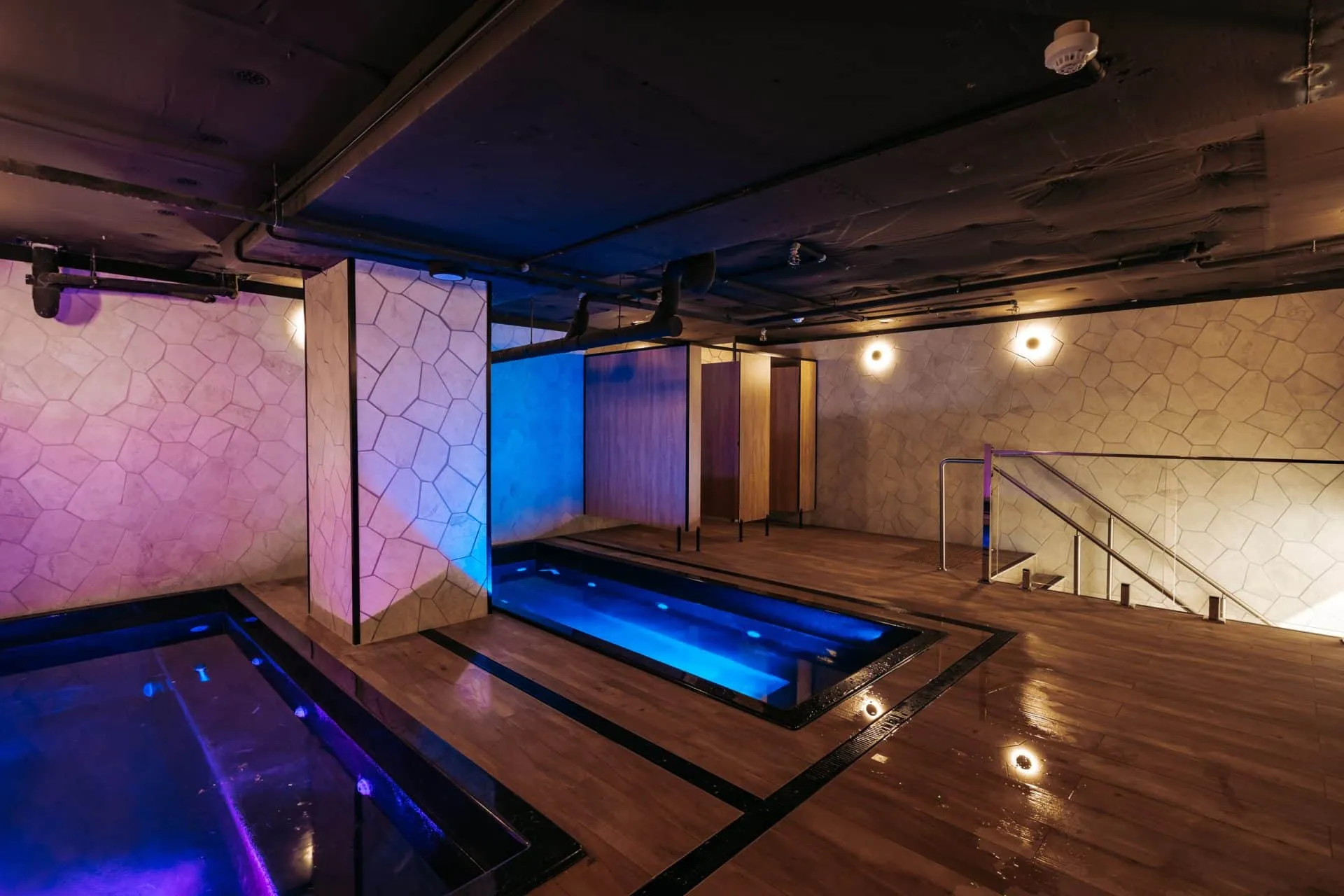 The recovery pool room from the far corner: one pool lit violet in the foreground and a second lit blue beyond a stone-clad pillar, on a timber deck, with a glass balustrade and stair to one side.