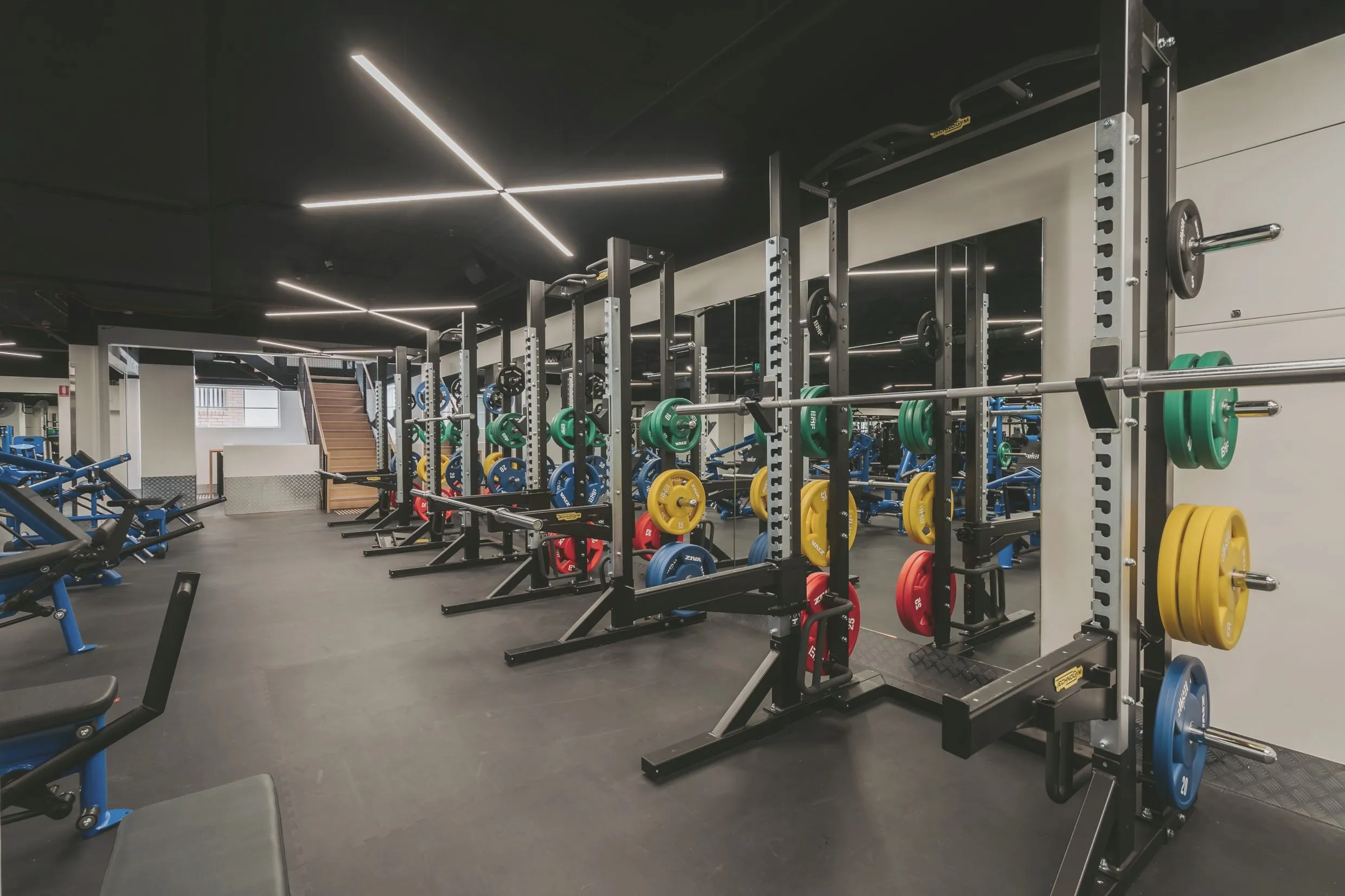 A line of half racks on the strength floor, barbells loaded with colour-coded plates, mirrors behind and a timber staircase beyond.